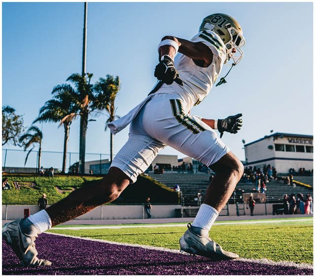 An adult wearing a football uniform and helmet is running on a field with palm trees and bleachers in the background under a clear blue sky. Description generated by AI