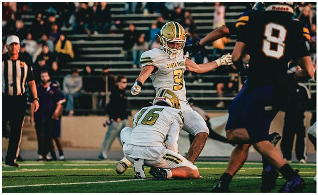 A group of people playing football on a field with one person kneeling and holding the ball while others are running nearby. Description generated by AI