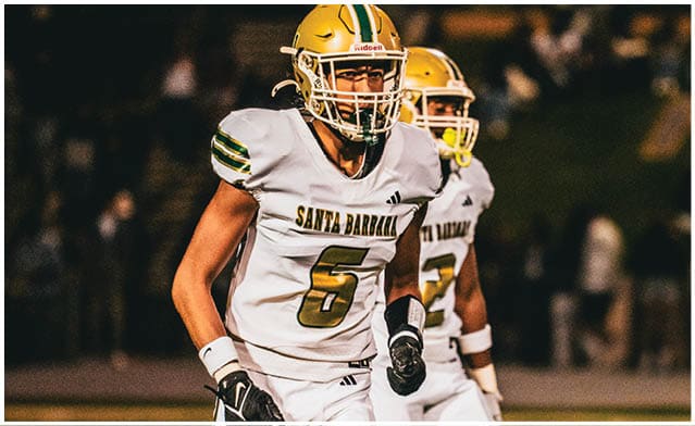 Two people wearing white football uniforms with yellow helmets are on a field, with one person in the foreground wearing a jersey labeled “Santa Barbara" and the number 6. Description generated by AI