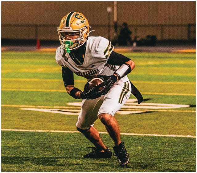 A person wearing a football uniform and helmet is crouched on a football field at night holding a football. Description generated by AI