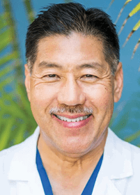 A smiling adult with short dark hair and a mustache is wearing a white coat over blue clothing with a blurred green and blue background. Description generated by AI
