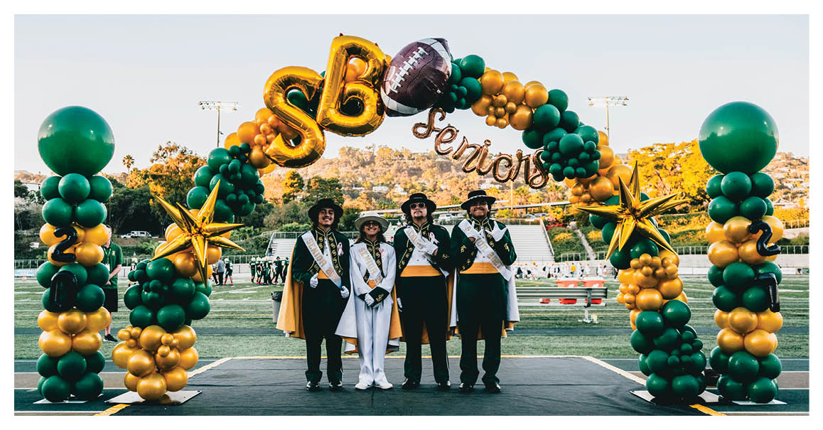 Four people dressed in formal attire with sashes and hats stand under a large balloon arch decorated with green and gold balloons, gold stars, and letters spelling “SBC Seniors" on a sports field. Description generated by AI