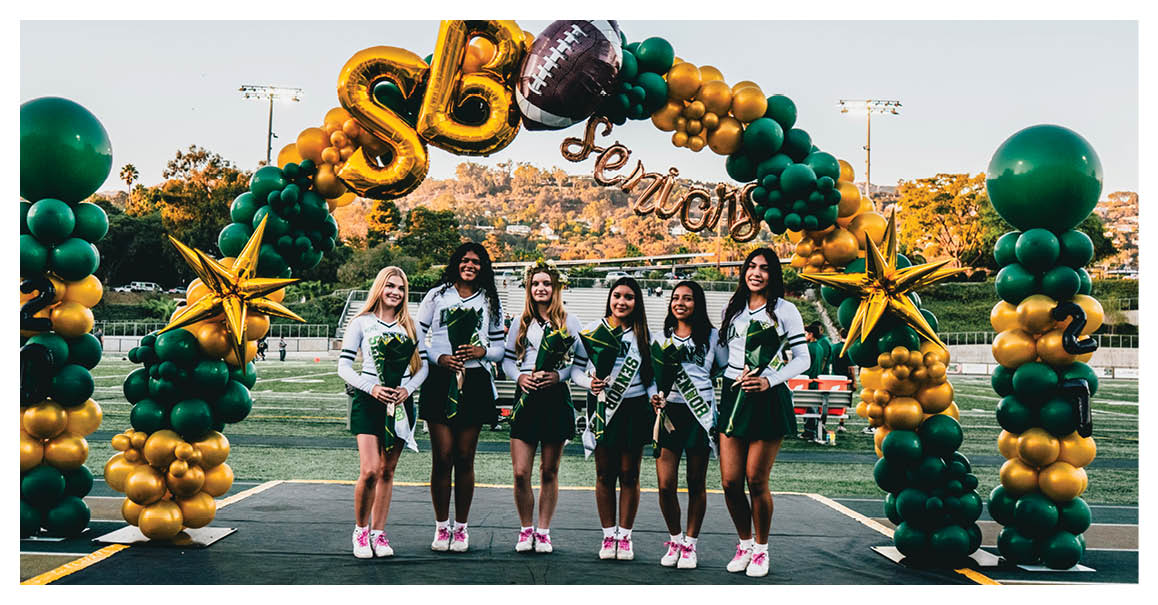 Six people wearing matching cheerleading uniforms stand under a green and gold balloon arch decorated with stars and the words “SB seniors" on a sports field. Description generated by AI