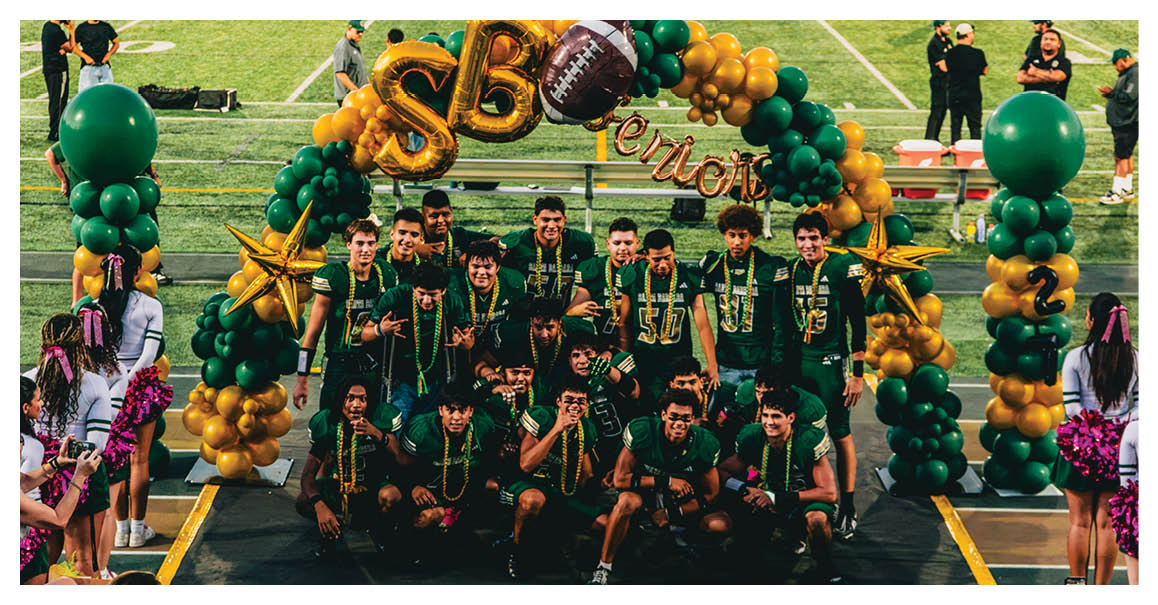 A group of people wearing matching sports jerseys pose together under a balloon arch decorated with a football and the word “Seniors" on a sports field with cheerleaders standing nearby. Description generated by AI