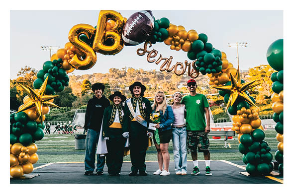 Six people stand together smiling under a green and gold balloon arch decorated with a football and the word seniors on a sports field. Description generated by AI