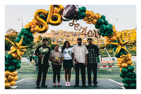 Five people stand together smiling under a green and gold balloon arch decorated with large gold stars and the words “SB Seniors" at an outdoor event on a sports field. Description generated by AI