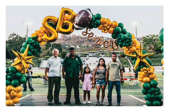 Five people stand together under a decorative balloon arch with gold and green balloons, large gold stars, a football balloon, and the word “Seniors" at an outdoor sports field. Description generated by AI