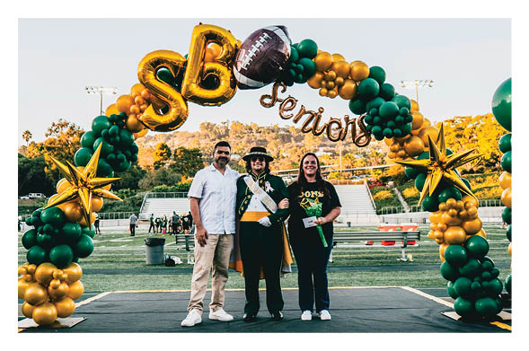 Three adults stand together on a sports field under a colorful balloon arch with “Seniors" written above, one wearing a sash and hat, smiling for a celebratory photo. Description generated by AI
