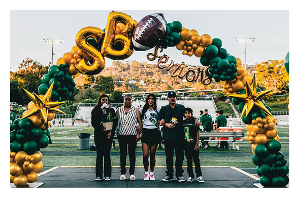 Five people stand together on a stage under a green and gold balloon arch decorated with stars and a football, with a sports field and trees in the background. Description generated by AI
