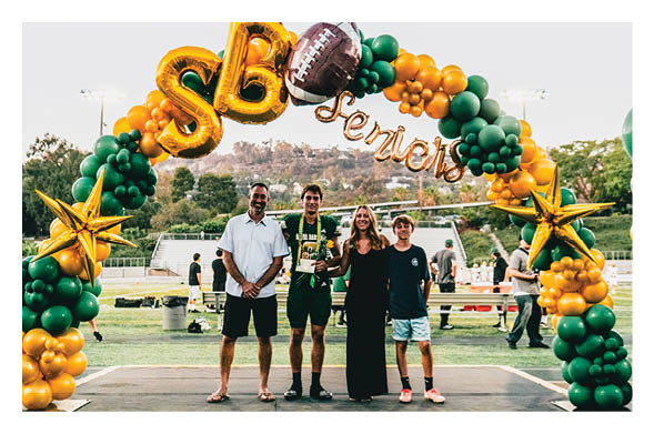 Four people stand smiling on a stage under a green and gold balloon arch decorated with stars and a football, with a sports field and hills in the background. Description generated by AI