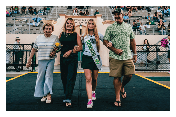 Four people walk arm in arm on a sports field with bleachers and spectators in the background, one wearing a sash that reads “Senior." Description generated by AI