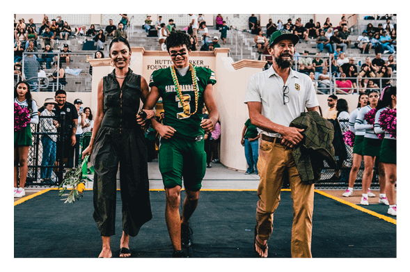 A person in a green sports uniform with face paint walks arm-in-arm with two adults on either side along a track with spectators and cheerleaders in the background. Description generated by AI