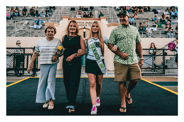 Four people walk arm in arm on a sports field with bleachers and spectators in the background, one wearing a sash that reads “Senior." Description generated by AI