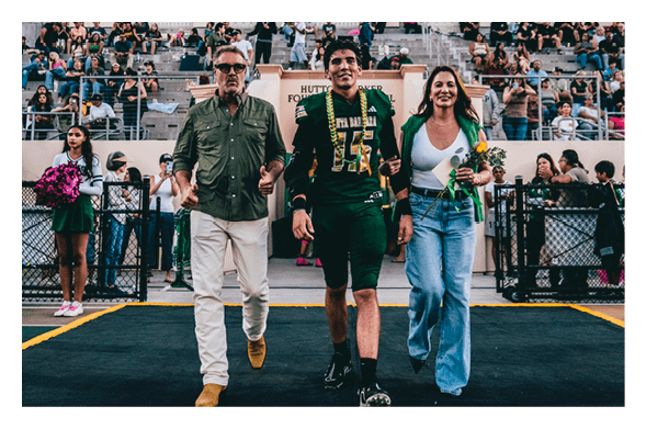 A person in a football uniform walks between two adults on a field with spectators in the background, while cheerleaders stand on either side holding pom-poms. Description generated by AI