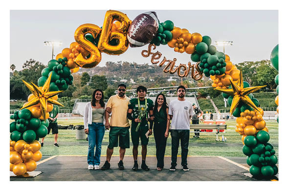 Five people stand together on a stage under a green and gold balloon arch with the word “Seniors" at an outdoor event on a sports field. Description generated by AI