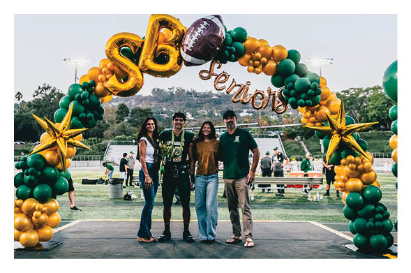 Four people stand smiling together under a decorative balloon arch on a sports field with bleachers and hills in the background. Description generated by AI