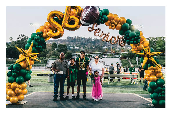 Five people, including two children and three adults, stand together on a stage under a green and gold balloon arch with the word “Seniors" in a sports field setting. Description generated by AI