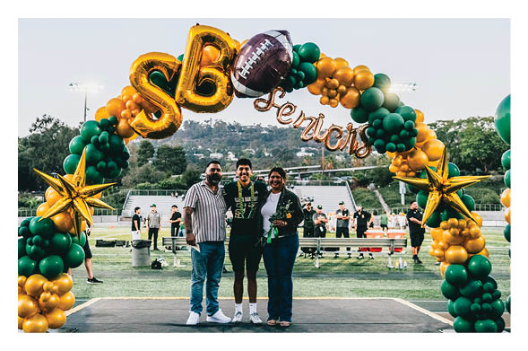 Three people stand together smiling on a stage decorated with green and gold balloon arches and star-shaped balloons at an outdoor event on a sports field. Description generated by AI