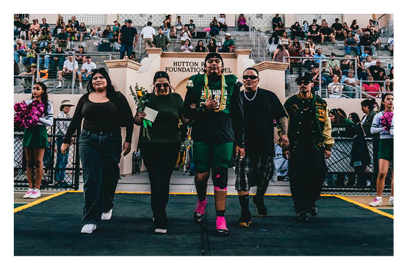 Five people walk confidently side by side on a track field with a crowd seated in bleachers behind them, one person holding flowers and another wearing a sports uniform with bright pink socks. Description generated by AI
