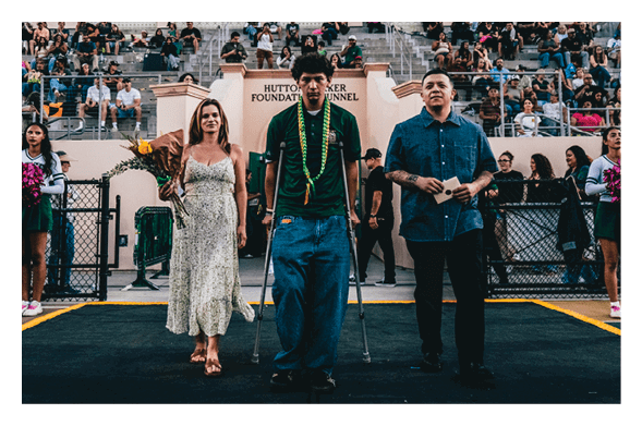 Three people stand on a stage in front of a seated crowd, with one person using crutches and wearing leis, another holding flowers, and a third holding an envelope. Description generated by AI