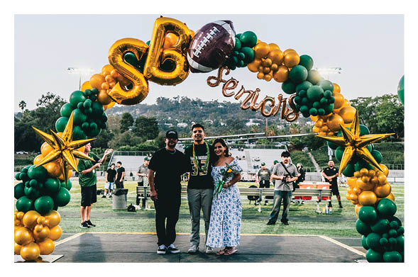 Three people stand together smiling under a green and gold balloon arch with the word “Seniors" at an outdoor event on a sports field. Description generated by AI