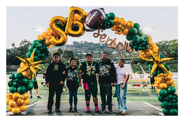 Five people stand together smiling under a decorative balloon arch with gold stars and green and gold balloons on a sports field during an outdoor event. Description generated by AI