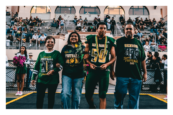 A group of four people wearing football-themed clothing walk arm in arm on a sports field with a crowd seated in the background. Description generated by AI