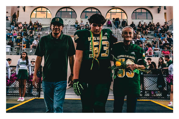 A person in a football uniform walks between two adults holding flowers on a field with spectators and cheerleaders in the background. Description generated by AI