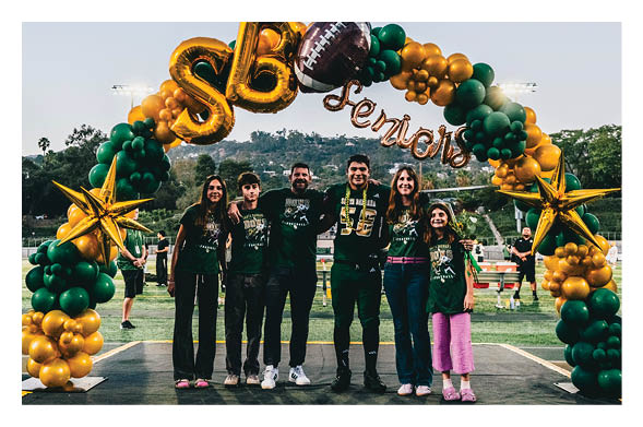Six people wearing matching dark green shirts stand closely together smiling under a balloon arch decorated with gold and green balloons and gold star-shaped balloons on a sports field. Description generated by AI