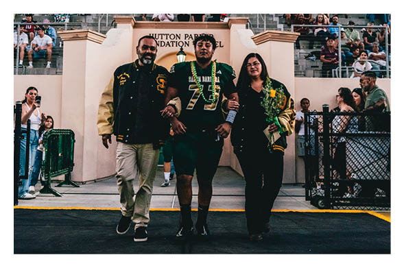 Three people walk arm in arm on a sports field with a crowd seated in bleachers behind them, one person wearing a sports jersey and the others dressed casually, while cheerleaders stand on either side. Description generated by AI