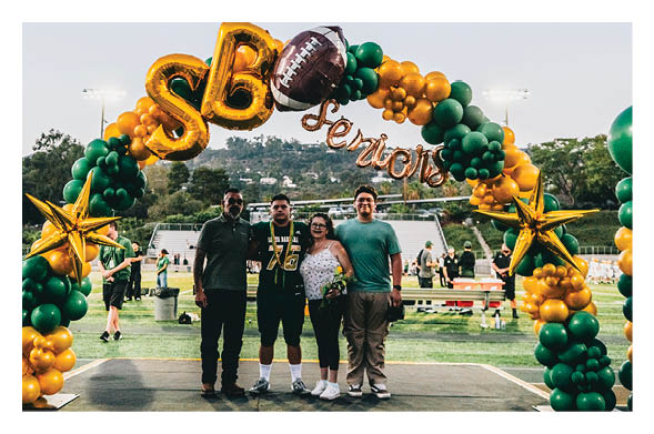 Four people stand together smiling under a green and gold balloon arch with the word “Seniors" at an outdoor sports field during a celebration. Description generated by AI