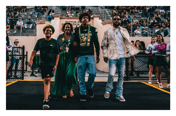 Four people walk arm in arm down a pathway in a stadium with spectators in the background, two wearing casual clothes and one in a long green dress holding flowers. Description generated by AI