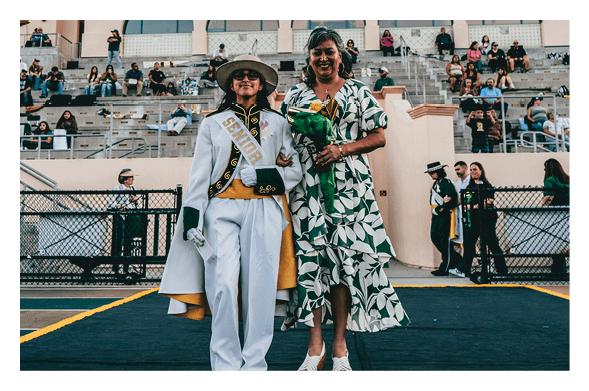 A person dressed in a white ceremonial outfit with a sash and hat stands smiling next to another person in a green and white patterned dress holding a bouquet, with spectators seated in bleachers behind them. Description generated by AI