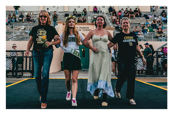 Four people linked arm-in-arm walk confidently on a stage in a stadium, wearing casual and cheer-themed clothing with spectators in the background. Description generated by AI