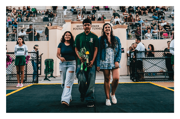 Three adults stand side by side on a track in front of a stadium crowd, with one holding flowers and two people in cheerleading uniforms visible nearby. Description generated by AI