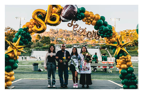 Four people stand smiling on a sports field under a green and gold balloon arch with “SB Seniors" balloons, with one person wearing a sash and holding flowers. Description generated by AI