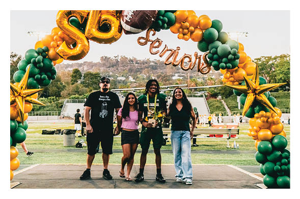 Four people stand smiling on a stage under a balloon arch decorated with gold stars and green and yellow balloons with the word “Seniors" and a football balloon above them at an outdoor sports field. Description generated by AI