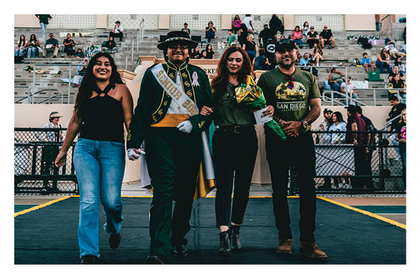 Four people walk arm in arm on a track in front of bleachers with seated spectators, with one person wearing a senior sash and holding a diploma. Description generated by AI
