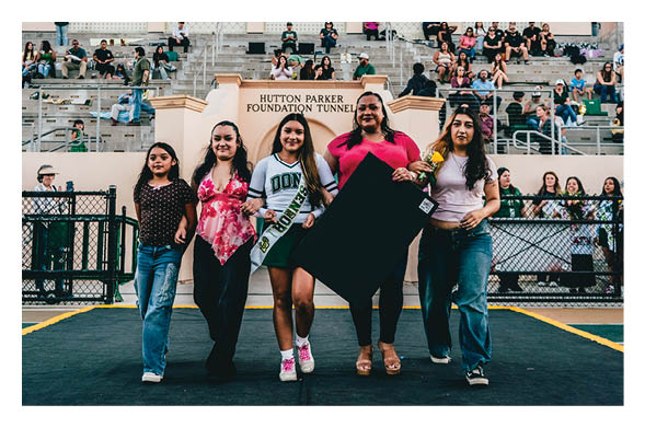 Five people standing confidently on a sports field in front of a stadium tunnel with spectators seated in the background, one holding a large black poster board. Description generated by AI