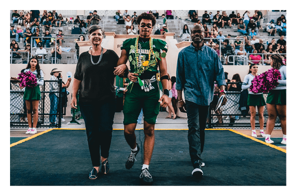 A person in a green sports uniform with a flower lei walks arm-in-arm with two adults on a sports field with spectators and cheerleaders in the background. Description generated by AI