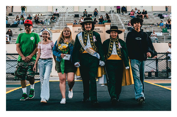 Five people walk arm in arm on a decorated stage with balloons and a crowd seated in bleachers behind them, two wearing ceremonial sashes and hats. Description generated by AI