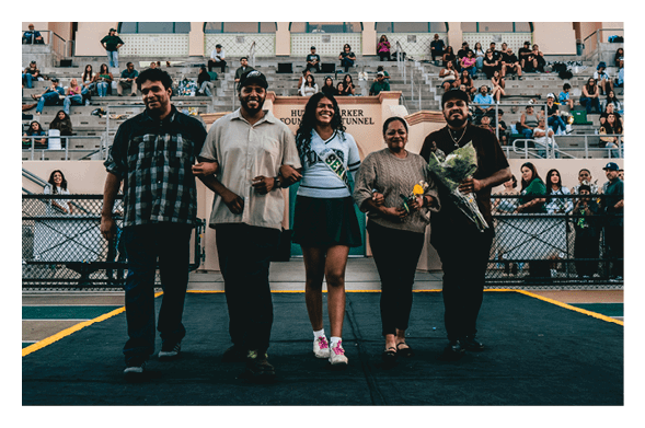 Five people stand arm in arm on a sports field with bleachers and spectators in the background, two holding flowers and one wearing a sash. Description generated by AI