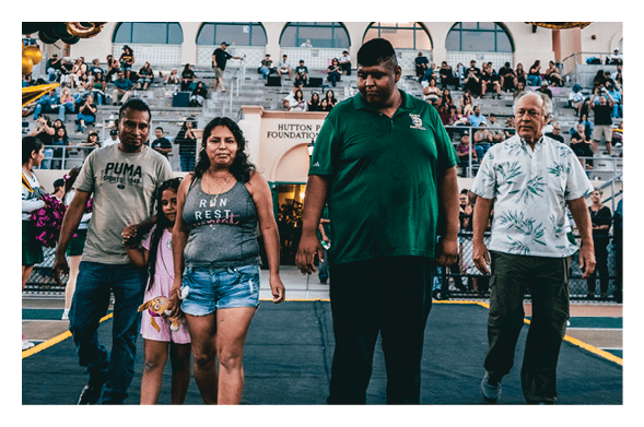 Five people, including one child holding a stuffed animal, walk together on a track in front of bleachers filled with seated people at an outdoor event. Description generated by AI