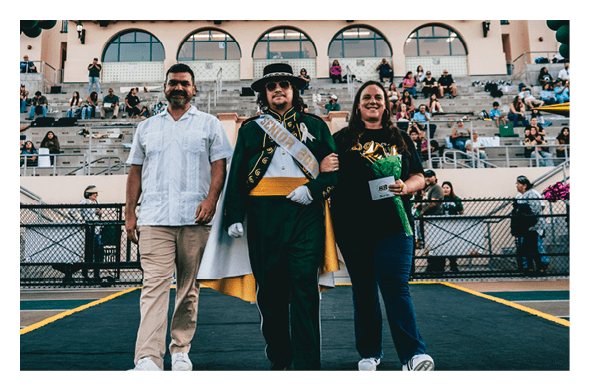 Three people stand arm in arm on a sports field with bleachers and spectators in the background, one person wearing a ceremonial cape, sash, and hat. Description generated by AI