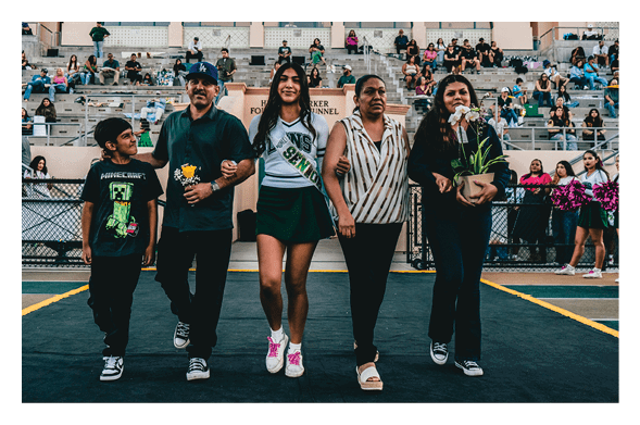 Five people walking arm in arm on a sports field with a crowd seated in bleachers behind them, one person wearing a cheerleading outfit and another holding a potted plant. Description generated by AI