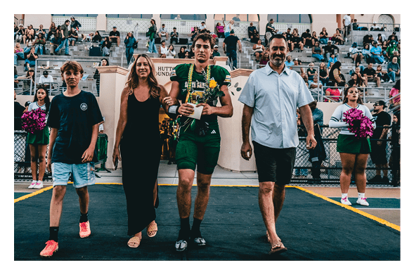 Four people walk arm in arm on a track with two people holding flowers, while cheerleaders stand on either side and a crowd watches from bleachers in the background. Description generated by AI