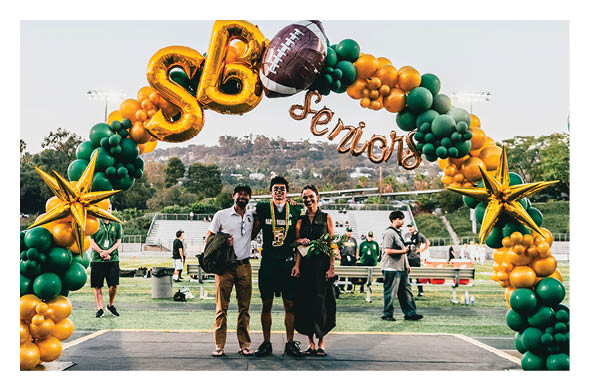 Three adults stand smiling on a sports field under a decorative balloon arch with gold stars, green and gold balloons, and a football balloon, celebrating a senior event. Description generated by AI