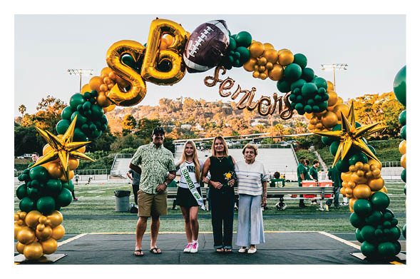 Four people stand together smiling under a green and gold balloon arch decorated with a football and the words “SB seniors" on a sports field. Description generated by AI