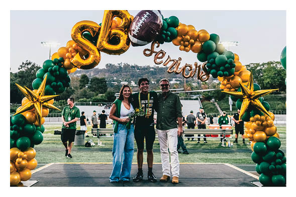 Three people stand smiling under a green and gold balloon arch decorated with stars, a football, and the word “Senior" on a sports field. Description generated by AI