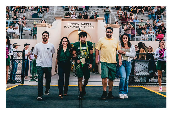 Five people walk arm in arm on a sports field with cheerleaders on either side and spectators seated in bleachers behind them. Description generated by AI